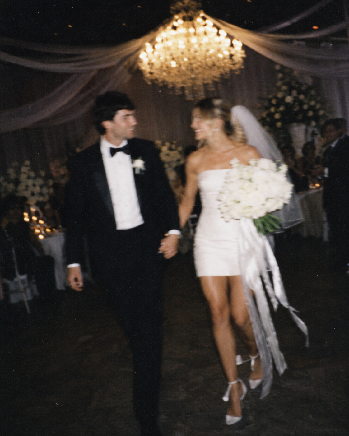 Bride and groom walking hand in hand through their wedding reception under a chandelier
